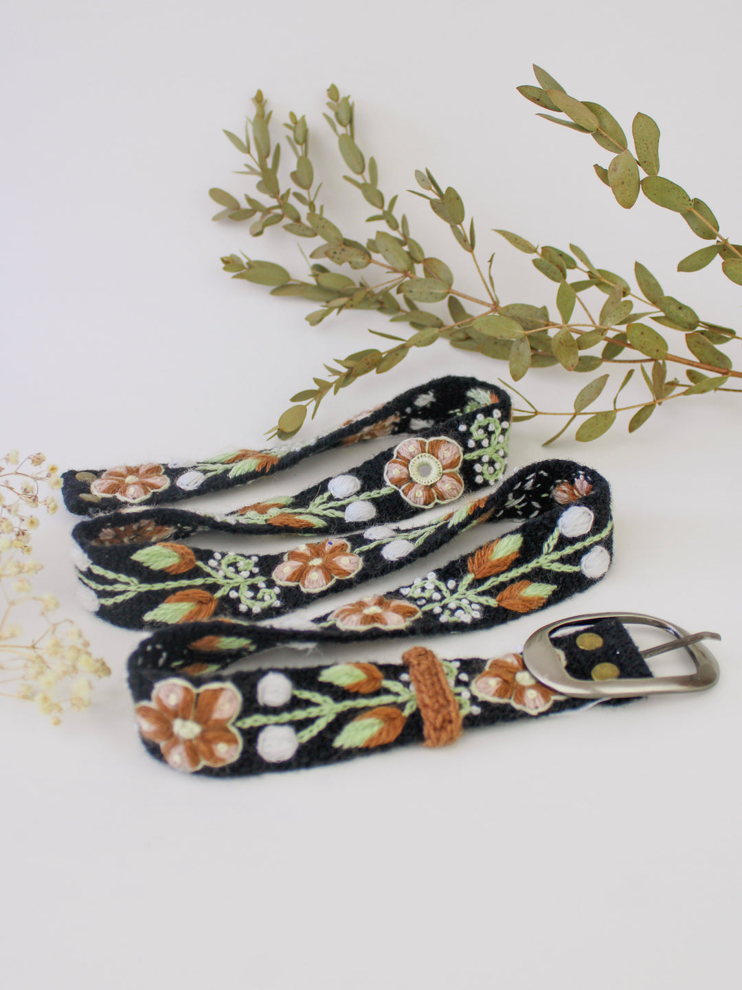 Embroidered black belt with brown, white, and green flowers and vines with a silver buckle on a white background with white flowers and green leaves in the background.