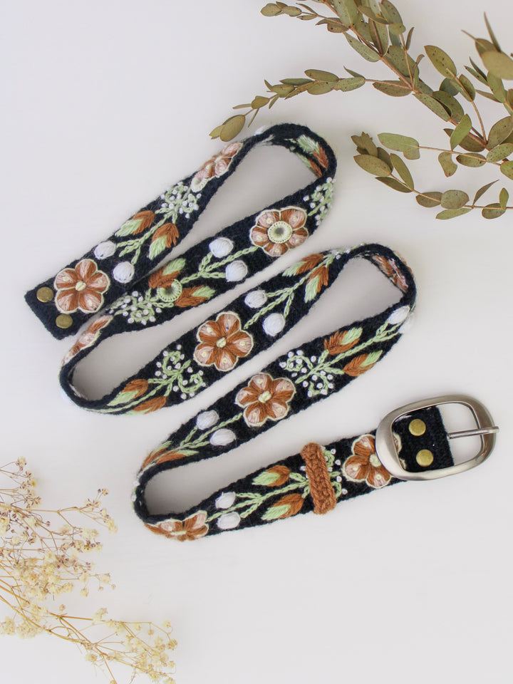 Embroidered black belt with brown, white, and green flowers and vines with a silver buckle on a white background with white flowers and green leaves in the background.