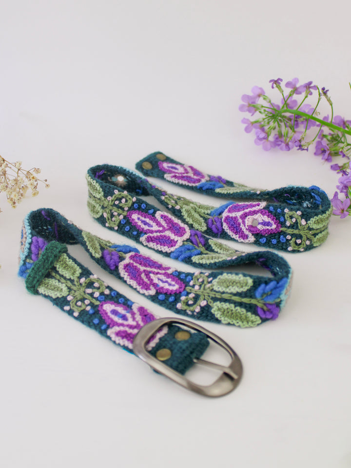 Embroidered bet with blues, greens, and purples, a silver buckle, on a white background with a sprig of white flowers and purple flowers.
