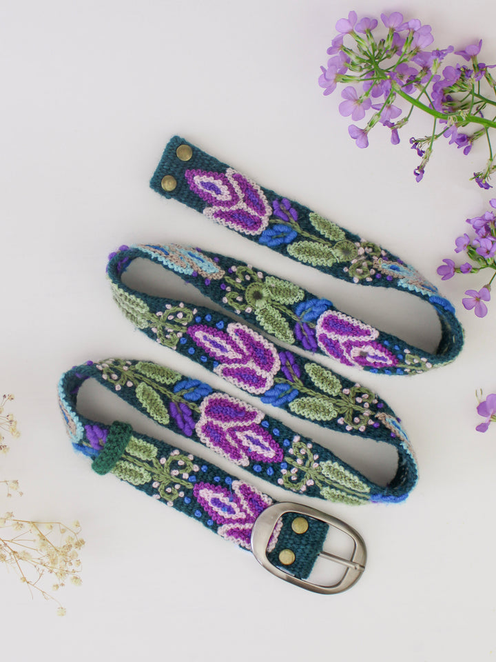 Embroidered bet with blues, greens, and purples, a silver buckle, on a white background with a sprig of white flowers and purple flowers.