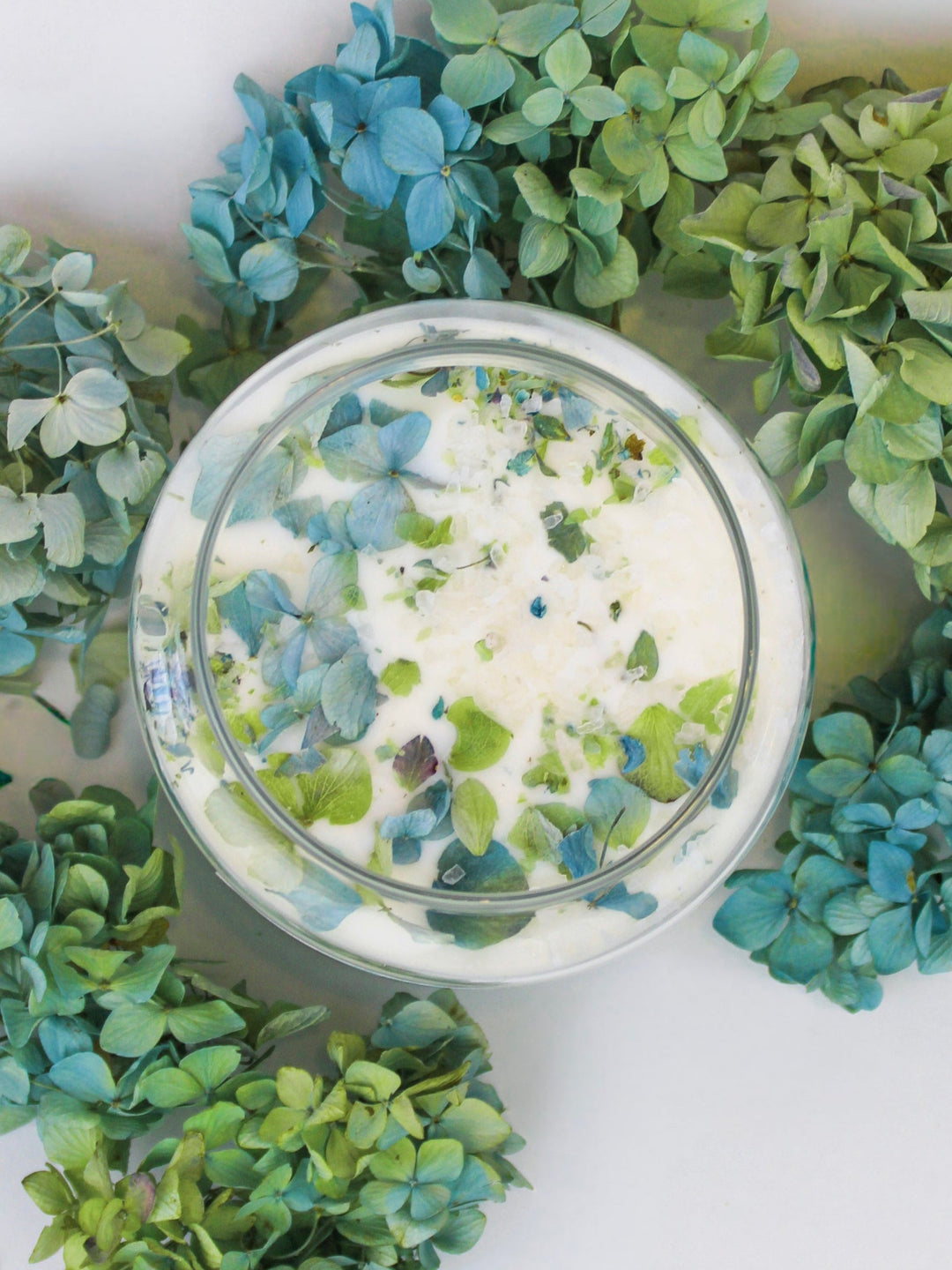 Glass soy wax candle with dried blue and green hydrangea petals on top.