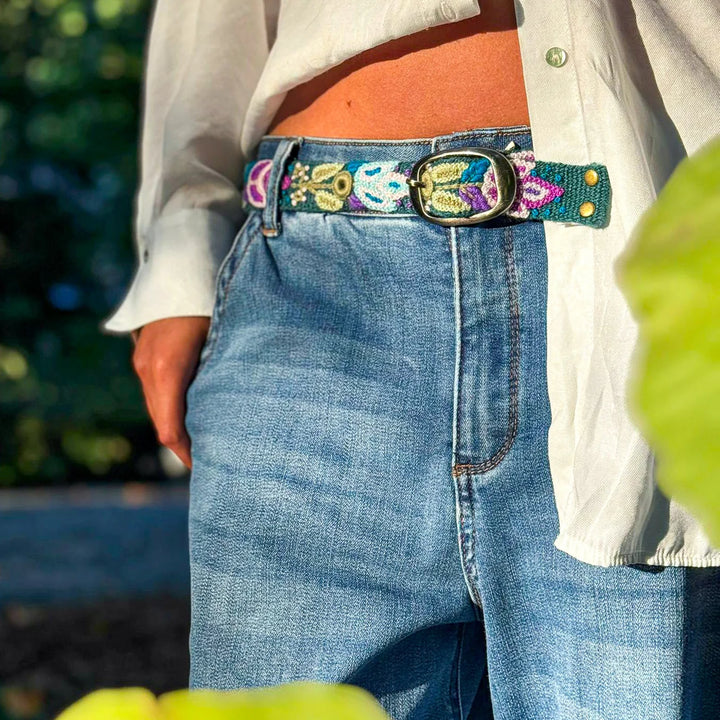 Embroidered bet with blues, greens, and purples, a silver buckle, on blue jeans .nd a white button down with green leaves in the background and foreground.