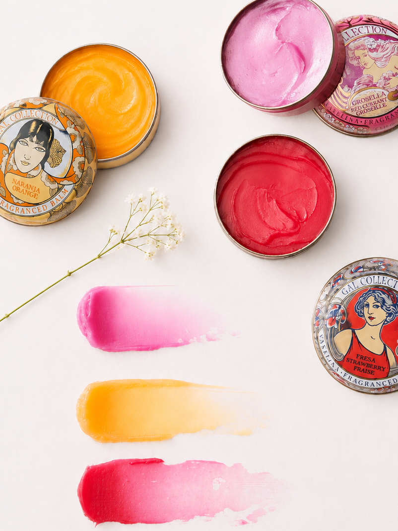 Four round metal tins of tinted lip balms arranged on a white background, with three product color smears below. The tins have art deco style illustrated lids and show orange, red currant, and strawberry fragrances.