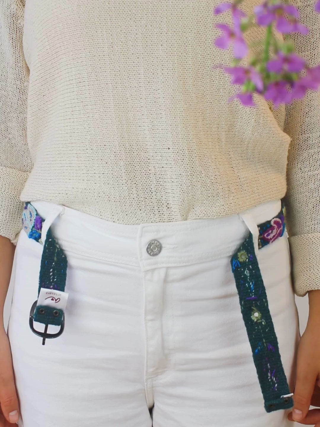 Embroidered bet with blues, greens, and purples, and a silver buckle being fastened over white pants and a beige sweater with purple flowers in the corner.