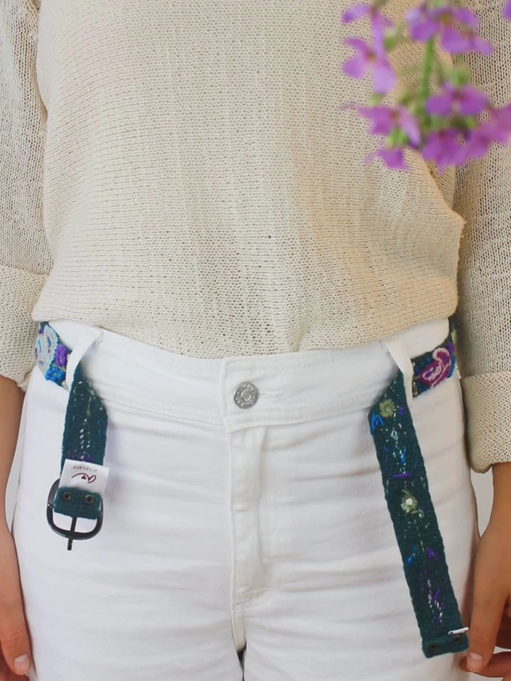 Embroidered bet with blues, greens, and purples, and a silver buckle being fastened over white pants and a beige sweater with purple flowers in the corner.
